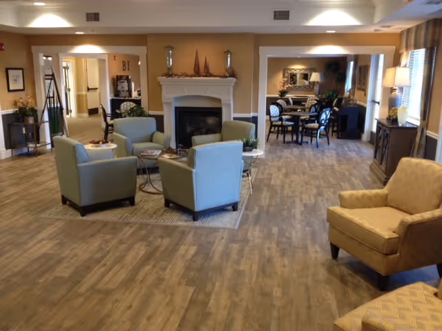 A spacious senior living lounge with armchairs arranged around a fireplace and a dining table area visible in the background.