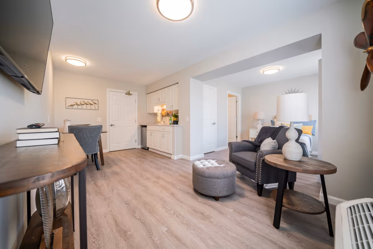 A bright and modern interior space featuring a small kitchen area with white cabinets and a countertop, a cozy living area with a gray armchair, a round wooden side table with a white lamp, and a gray ottoman. In the background, a bedroom with a bed, pillows, and a nightstand with a lamp is visible. The room has light wood flooring and neutral-colored walls with ceiling lights.