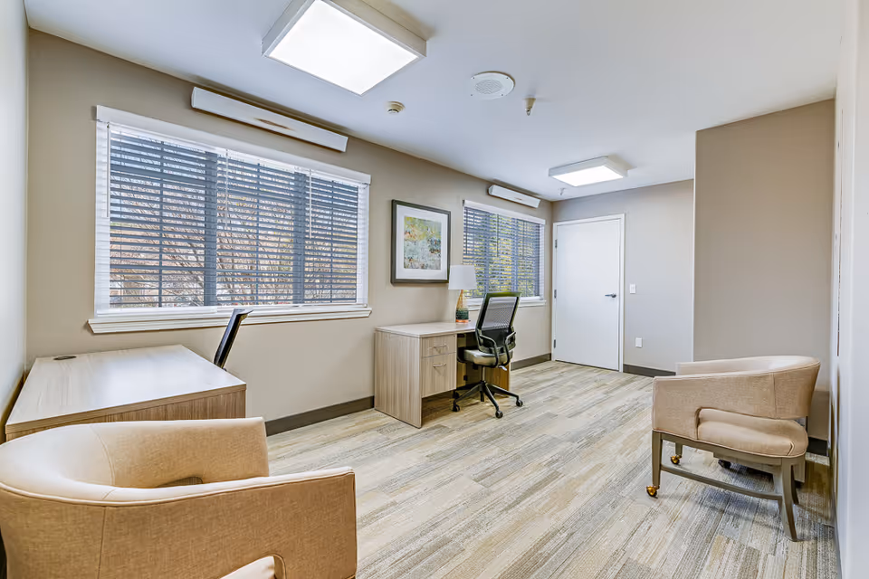 Well-lit interior room with two desks, an office chair, two armchairs, large windows with blinds, and neutral decor.
