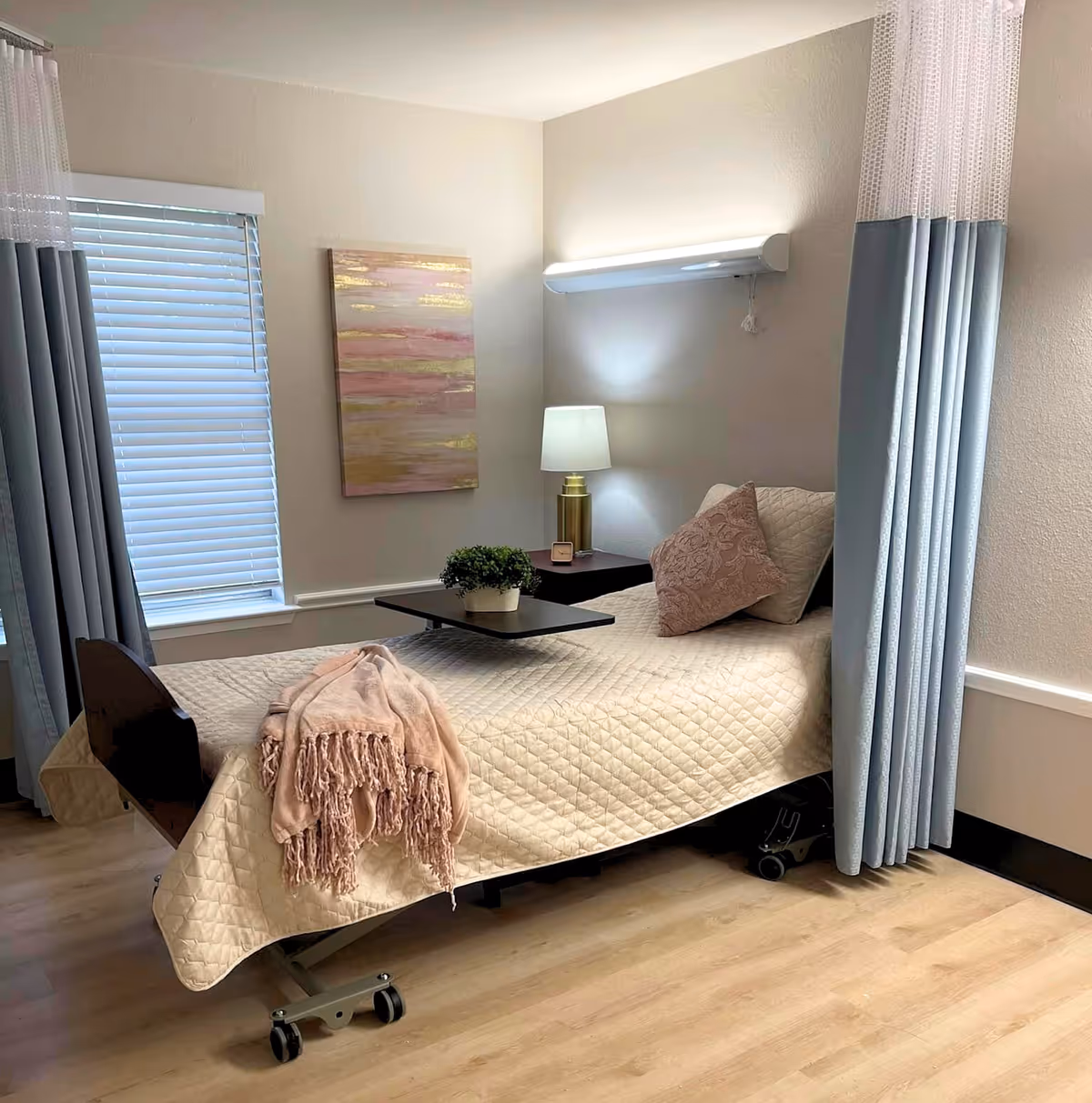 A neatly made hospital-style bed with a beige quilted blanket and a pink throw blanket draped over the foot. There is a pink decorative pillow and a small potted plant on an overbed table. The room has light-colored walls, a window with closed blinds and blue curtains, a bedside table with a lamp, and a piece of abstract wall art.