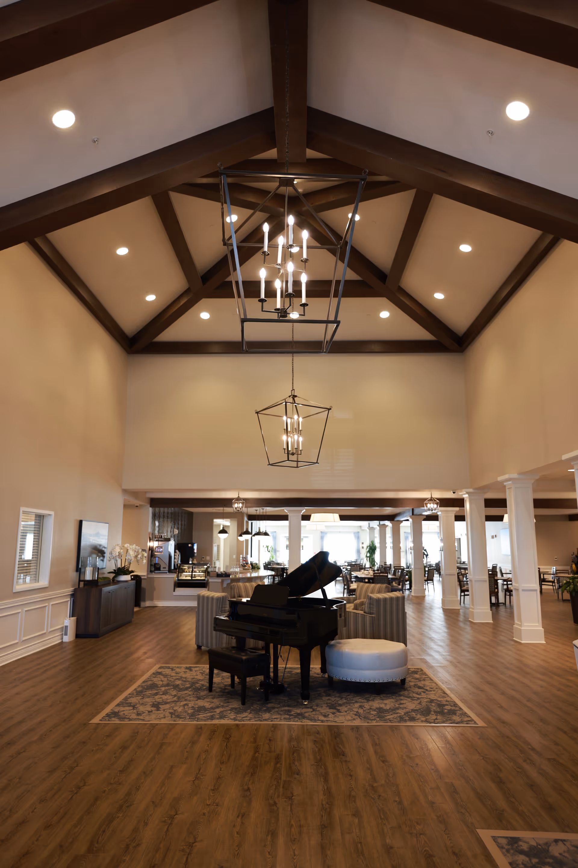 Spacious communal lounge with a grand piano on a rug, seating areas, and a high beamed ceiling with chandeliers.