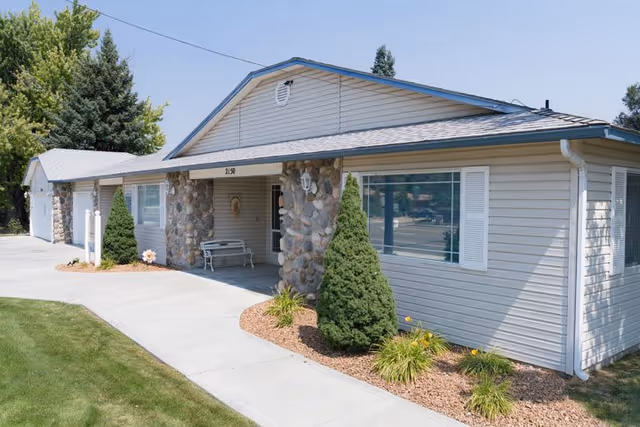 Single-story building with beige siding and stone pillars at the entrance, surrounded by small landscaped bushes and a concrete walkway, under a clear blue sky.