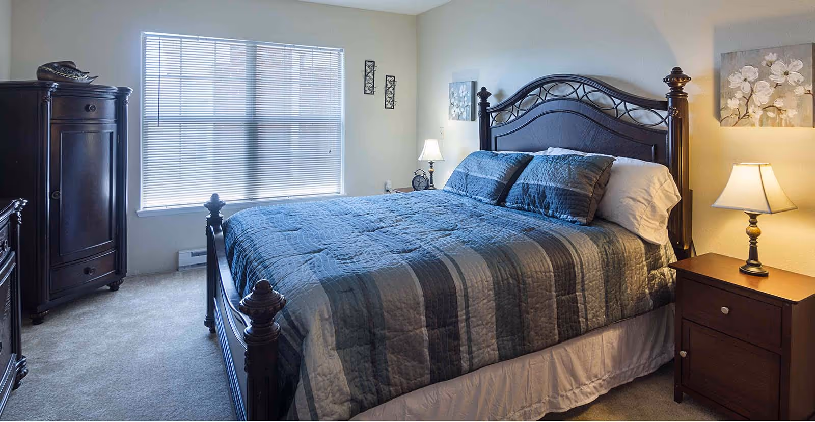A bedroom with a large dark wooden bed featuring a blue and gray striped quilt and matching pillows. There is a wooden nightstand with a lamp on the right side of the bed and a wooden armoire on the left side near a window with closed blinds. The walls are light-colored and decorated with floral artwork and small wall hangings.