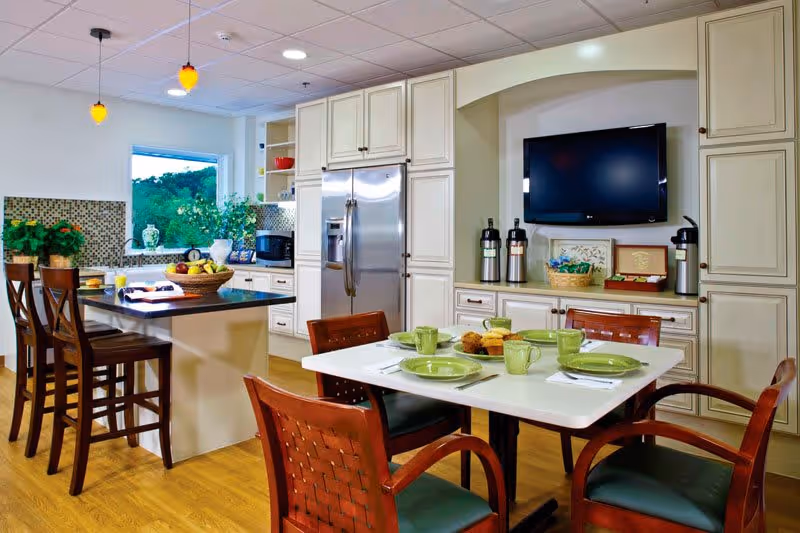 A bright and modern kitchen and dining area with white cabinetry, a stainless steel refrigerator, and a kitchen island with three wooden bar stools. The dining table is set with green plates, cups, and napkins, and there are four wooden chairs around it. A flat-screen TV is mounted on the wall above a countertop with coffee dispensers and a basket. There is a window with a view of greenery outside.