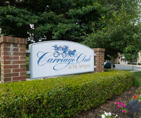 White entrance sign with a blue carriage logo reading "Carriage Club at Mt. Arlington" between brick pillars amid landscaped hedges and trees.