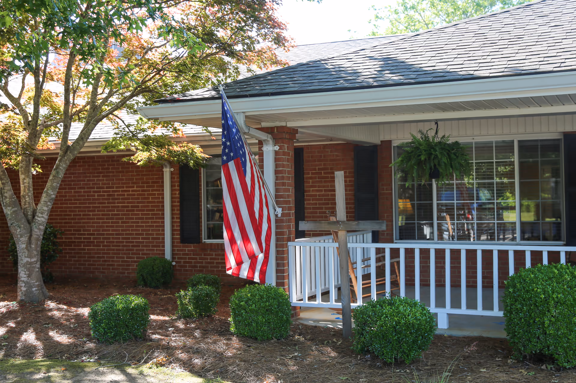 Front porch of a brick building with an American flag, white railing, hanging fern, and trimmed bushes.