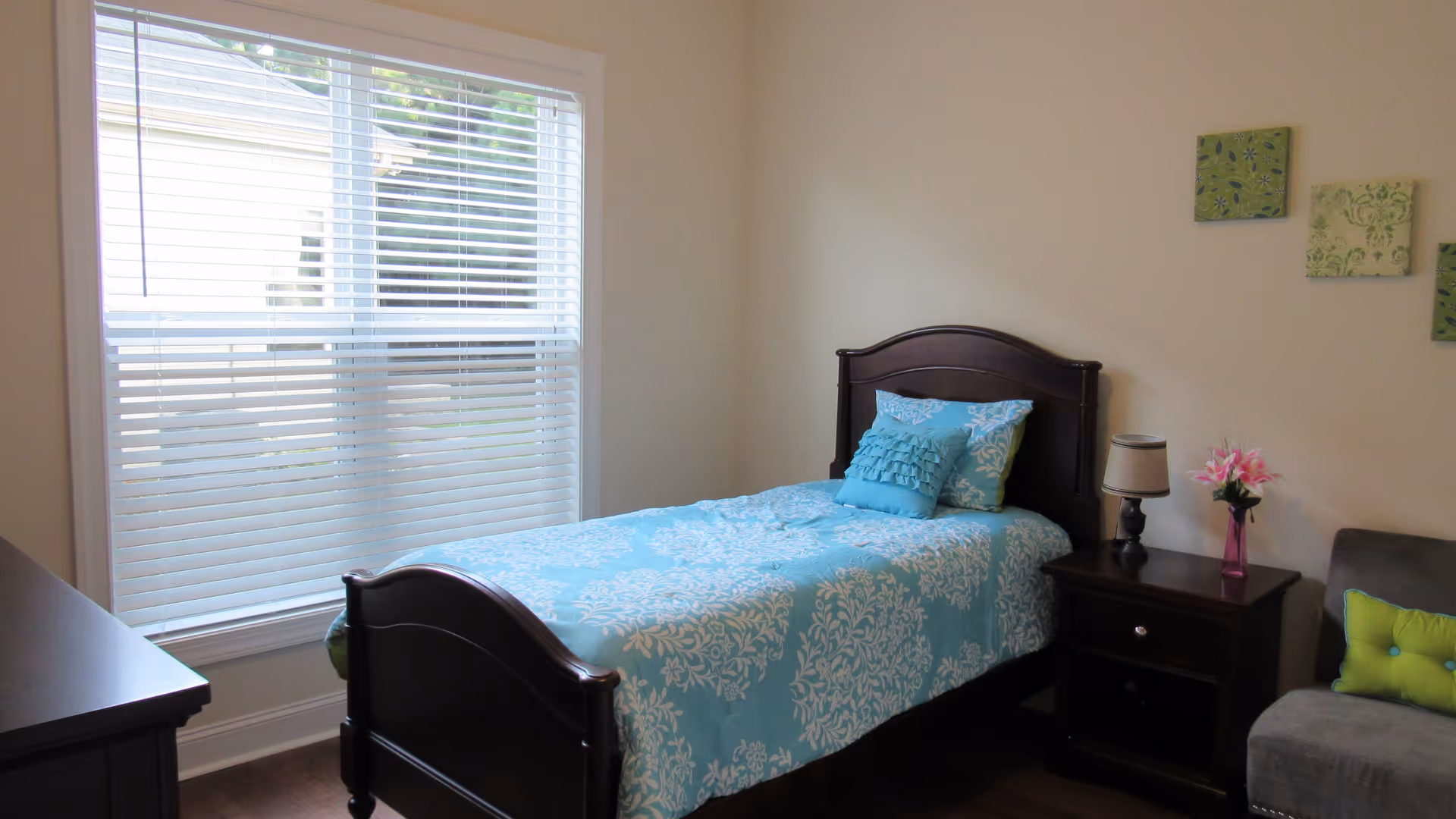 A bedroom with a single dark wood bed featuring blue and white patterned bedding and two decorative pillows. Next to the bed is a dark wood nightstand with a small lamp and a pink vase with flowers. A gray chair with a green pillow is partially visible on the right. The room has a large window with white blinds and light-colored walls decorated with three small square art pieces.