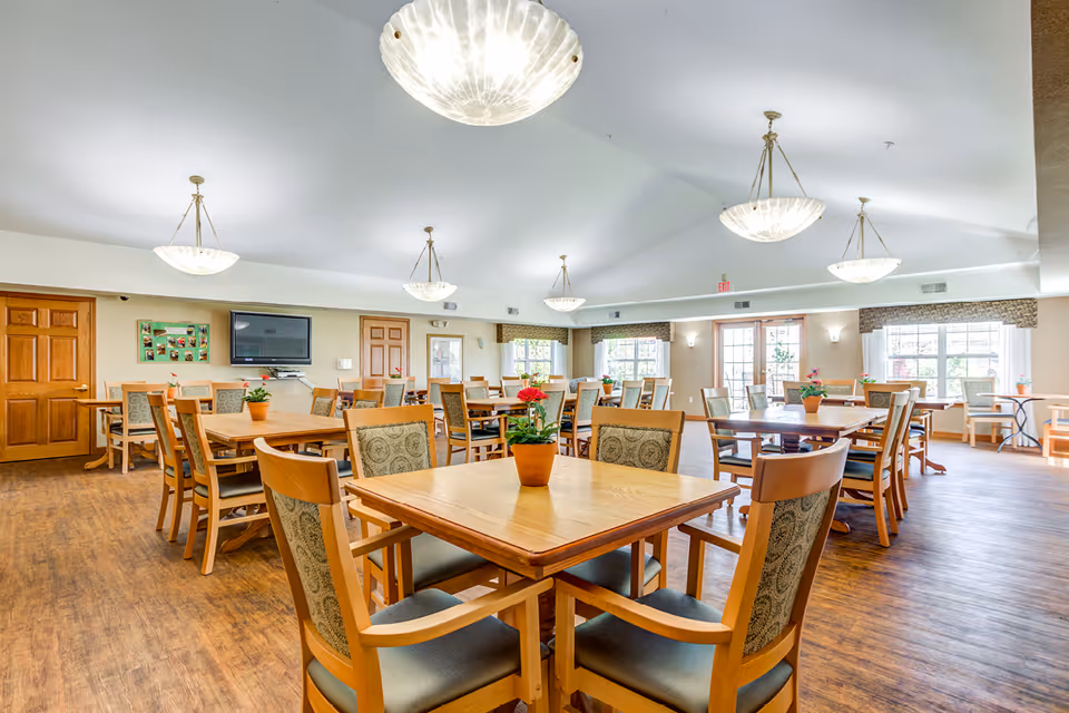 A spacious dining room with multiple wooden tables and chairs arranged neatly. Each table has a small potted plant with red flowers. The room has large windows allowing natural light to fill the space, and several ceiling lights provide additional illumination. The floor is wooden, and there is a wall-mounted television and bulletin board on one side.