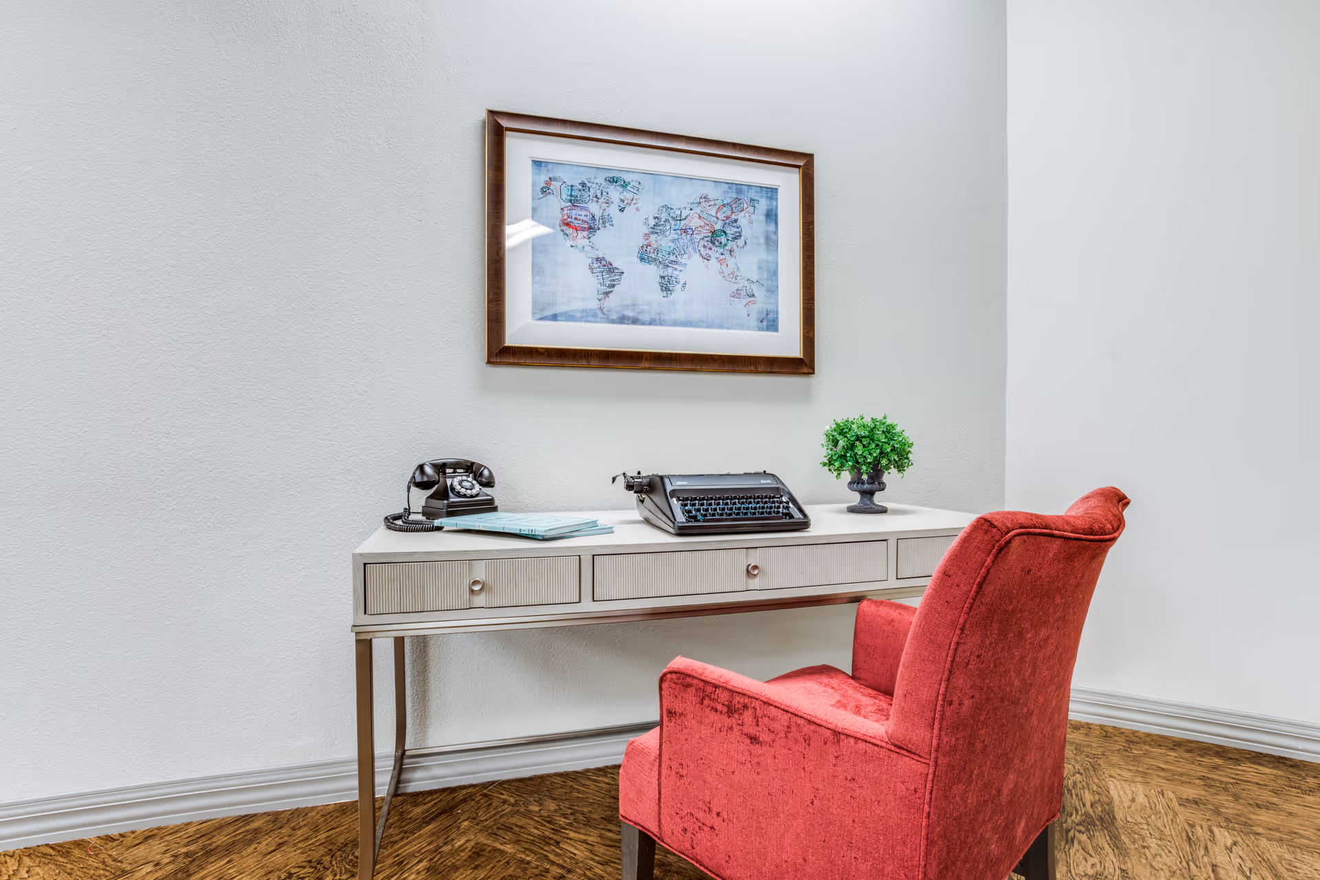 A small desk with a vintage typewriter and telephone beneath a framed world map, with a red upholstered chair in front.