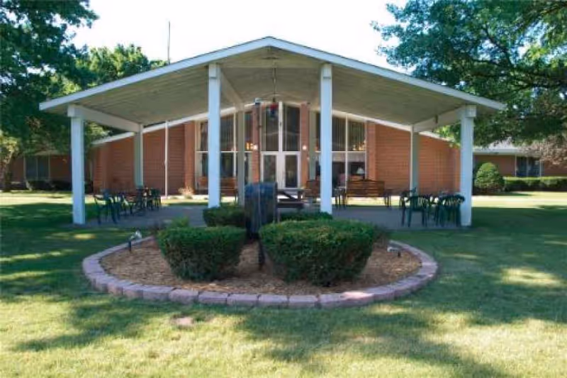 Covered outdoor patio with seating and a circular landscaped planter in front of a low brick building.