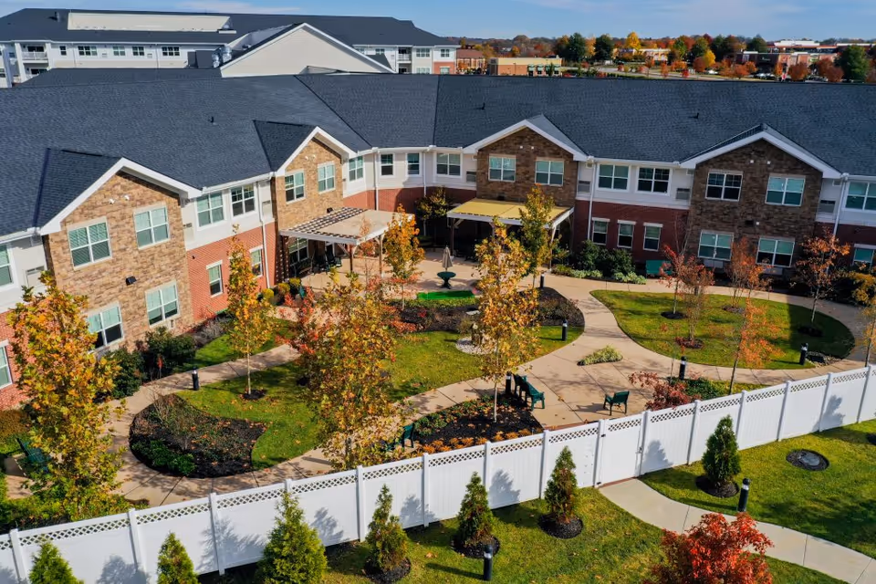 Aerial view of a senior living facility courtyard with a paved walking path, green lawn, trees with autumn foliage, benches, and a white fence surrounding the area. The building has a brick and stone exterior with multiple windows and a dark roof.
