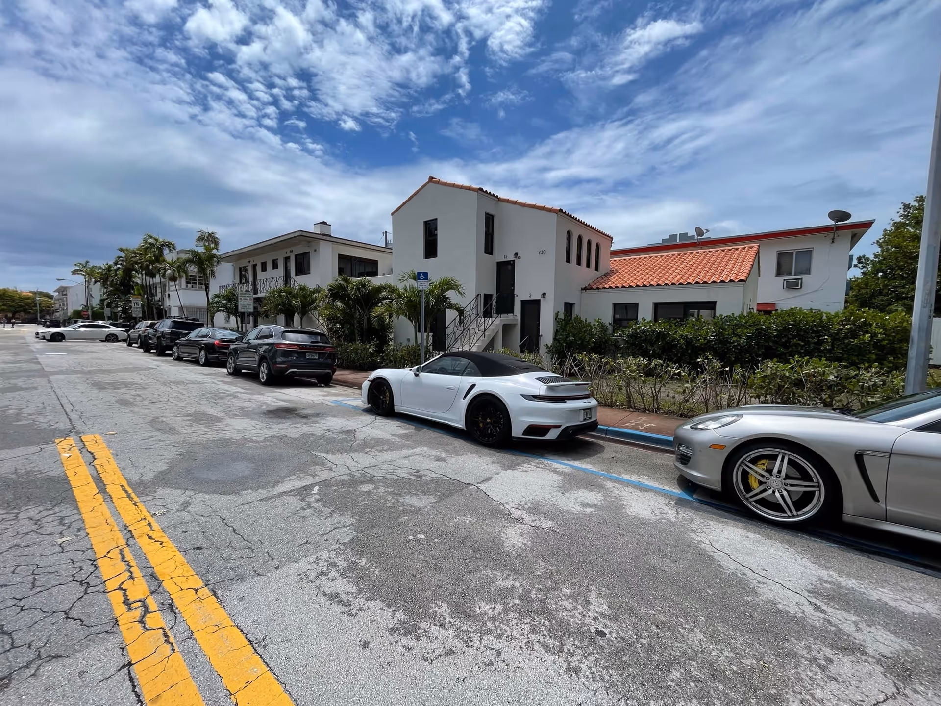 Street view of a white Mediterranean-style building with a red tile roof and several cars parked along a cracked road.
