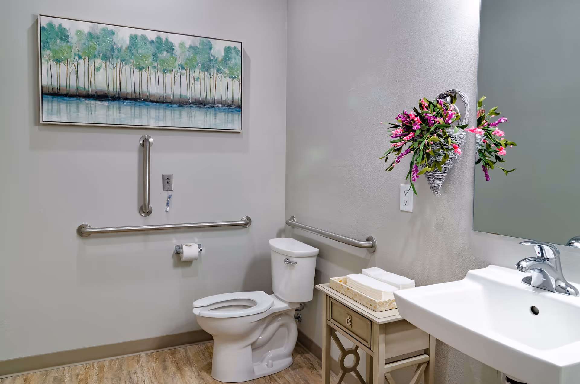 A clean and accessible bathroom featuring a white toilet with grab bars on the walls, a white sink with a chrome faucet, a small side table holding a tissue box and folded towels, a wall-mounted mirror, a decorative heart-shaped basket with pink and purple flowers, and a framed painting of trees above the toilet.