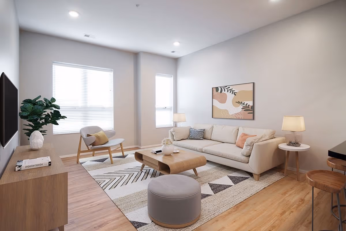 A modern living room with light-colored walls and wooden flooring. The room features a beige sofa with multiple cushions, a wooden coffee table, a round gray ottoman, a wooden side table with a lamp, a wooden chair with a yellow cushion, a large patterned rug, a flat-screen TV mounted on the wall, and a wooden TV stand with a plant and a book on it. Two windows with blinds allow natural light into the room, and a framed abstract artwork hangs above the sofa.