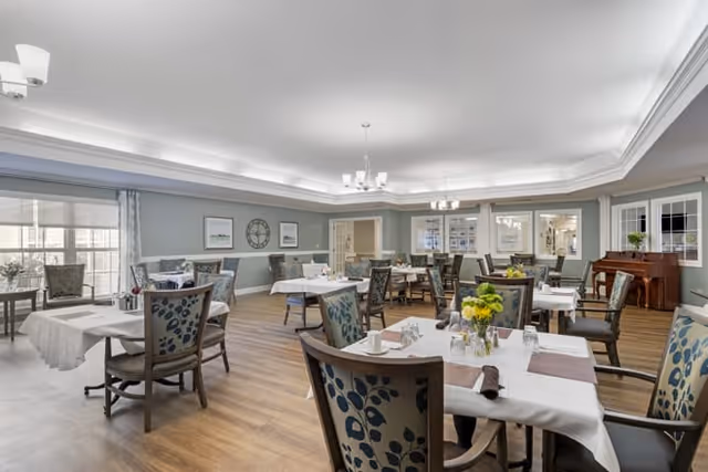A spacious dining room with multiple tables covered in white tablecloths, each set with glasses, napkins, and small flower arrangements. The room has wooden flooring, light blue walls with white trim, large windows letting in natural light, and a piano in the corner. Comfortable chairs with patterned upholstery surround the tables.