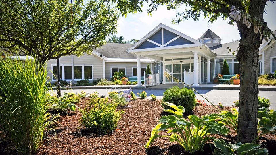 Front exterior view of Havenwood Heritage Heights facility with a landscaped garden featuring various green plants and trees in the foreground, and a covered entrance with white pillars and large windows in the background under a clear sky.