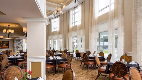 Spacious double-height dining room with round tables set with black tablecloths and chairs, tall windows with sheer curtains, and chandeliers.