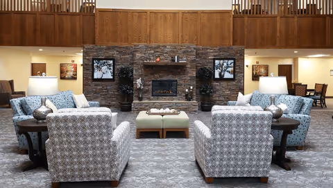 A spacious communal lounge with patterned armchairs and sofas arranged around a central stone fireplace, ottomans, table lamps, and a wooden balcony above.