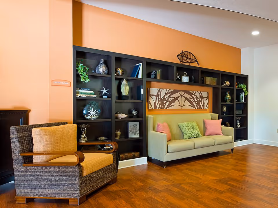 A cozy living room area with a beige sofa adorned with green and pink pillows, a cushioned armchair with wooden armrests, and a large black shelving unit filled with decorative items such as books, vases, and sculptures. The walls are painted in a warm orange and white color scheme, and the floor is covered with wood-style flooring.