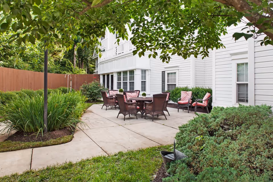 Outdoor patio area with several wicker chairs and tables arranged on a concrete surface next to a white building. The patio is surrounded by green bushes, plants, and trees, with a wooden fence in the background.