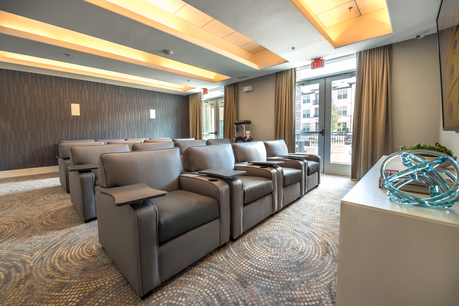 A modern seating area with multiple rows of gray recliner chairs equipped with small attached tables, arranged in a room with patterned carpet and a textured dark accent wall. The room has large glass doors with curtains leading outside, and a white cabinet with decorative items on the right side.