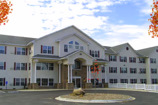 Front exterior view of a large, three-story senior living facility building with beige siding, multiple windows with red shutters, and a covered entrance supported by columns. There is a circular driveway with a landscaped island featuring a small tree and rocks. The sky is partly cloudy.