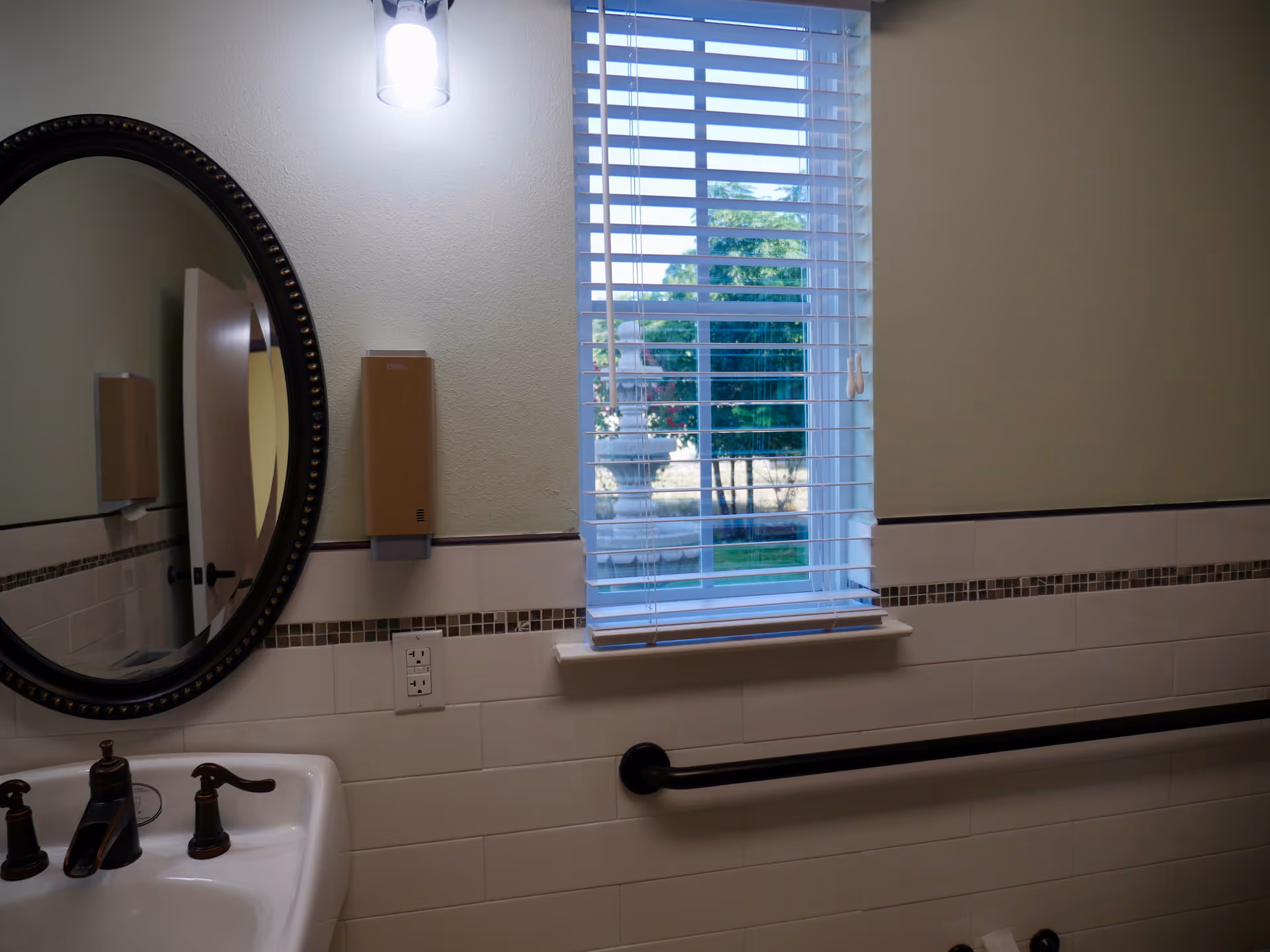 Bathroom interior with a sink, oval mirror, soap dispenser, grab bar, and a window with blinds showing trees outside.