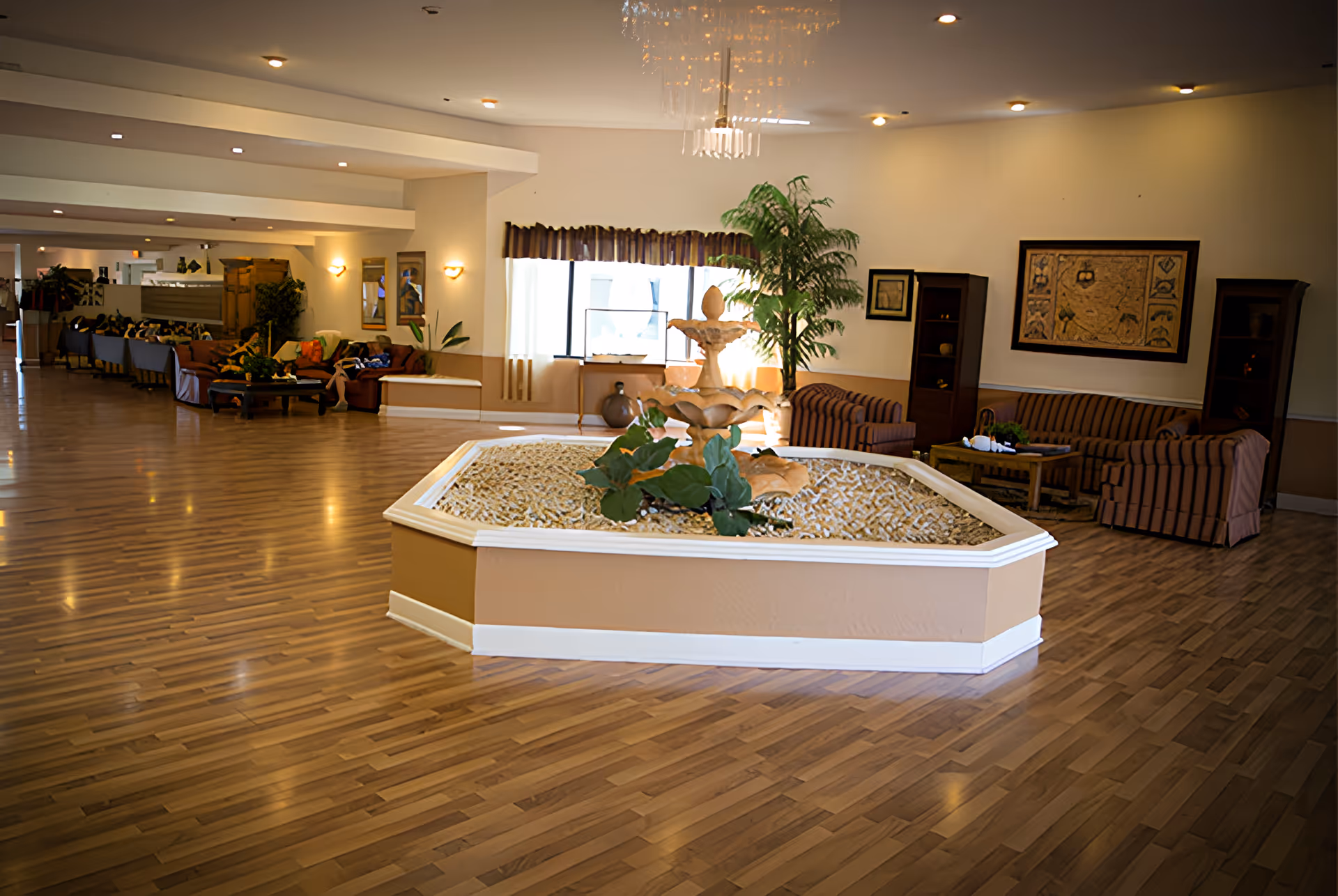 Spacious interior common area with wooden flooring, a central decorative fountain surrounded by pebbles and plants, striped sofas, a coffee table, framed artwork on the walls, and large windows with curtains allowing natural light.