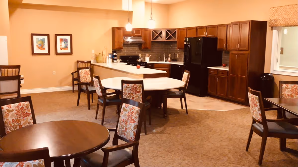 A cozy senior living facility common area featuring a kitchen with dark wooden cabinets, a black refrigerator, and a stove. The kitchen is adjacent to a dining area with several round and rectangular tables surrounded by chairs with floral patterned upholstery. The walls are painted a warm beige color, and two framed artworks hang on one wall. The space is softly lit with hanging pendant lights above the kitchen counter.