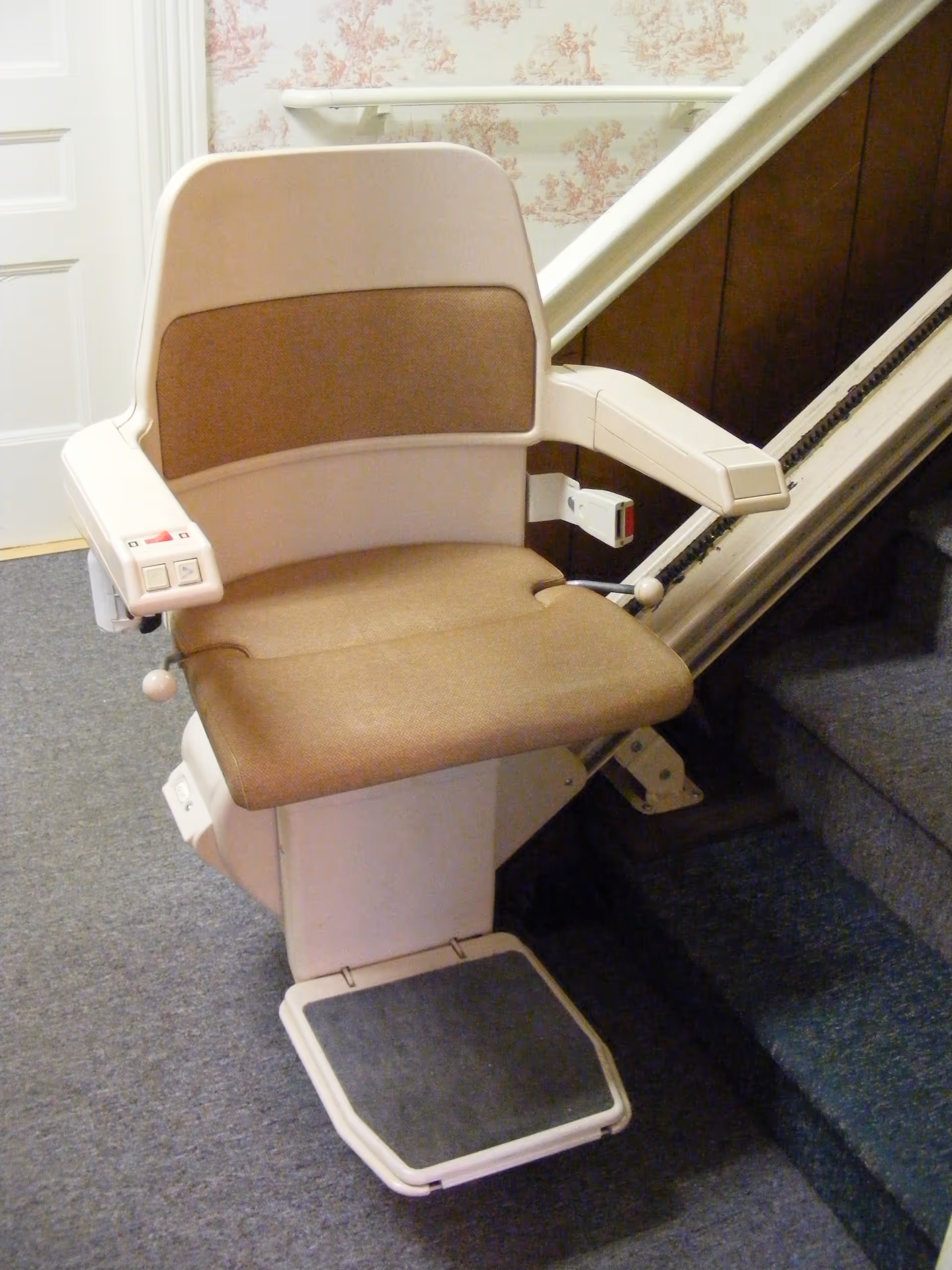 A beige and brown stair lift chair installed on a staircase with carpeted steps and floral wallpaper on the wall.