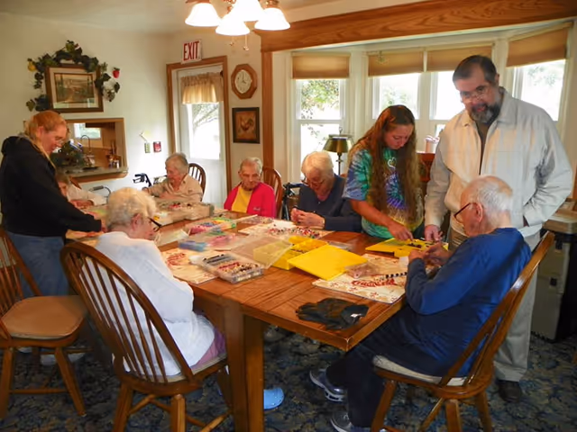 A group of elderly people and two caregivers are gathered around a wooden table in a well-lit room, engaging in arts and crafts activities with various colorful materials spread out on the table. The room has large windows with blinds, a clock on the wall, and a cozy, home-like atmosphere.