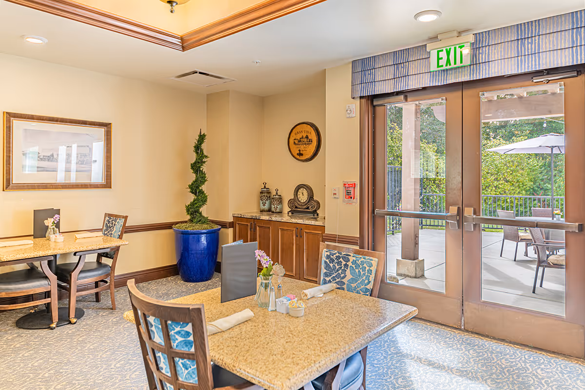 A bright dining area in a senior living facility with tables and chairs set for meals. The room features a large glass door leading to an outdoor patio with seating and an umbrella. Inside, there is a blue potted plant, a framed picture on the wall, and a cabinet with decorative items including a clock and jars. The exit sign is visible above the door.