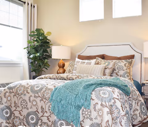 A cozy bedroom with a bed featuring a floral patterned comforter and multiple pillows. A teal knitted throw blanket is draped over the bed. Next to the bed is a wooden nightstand with a lamp that has a white shade and a wooden base. A large green potted plant is placed near a window with white blinds and beige curtains.