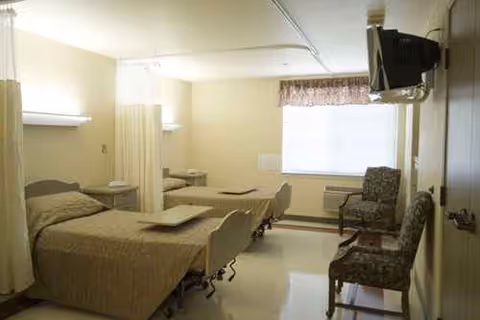 Two beds separated by a partition in a cream-colored patient room with chairs near a window.