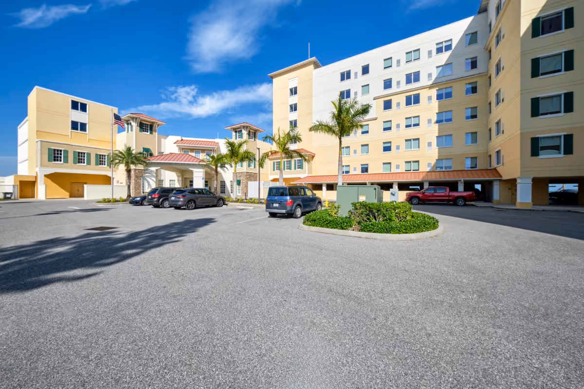 Exterior view of a multi-story senior living facility with a parking lot in front. The building is painted in light yellow and white with several windows and palm trees near the entrance. Several cars are parked in the lot under a clear blue sky.