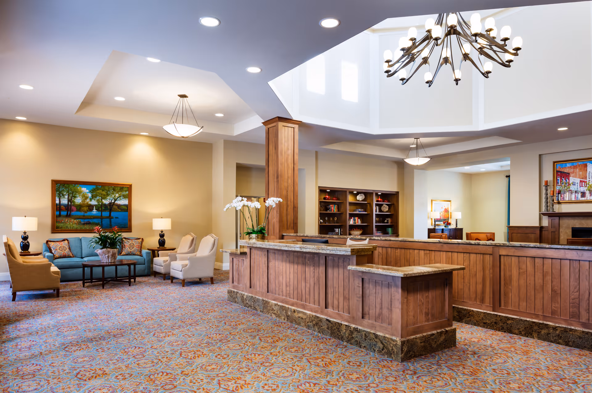 A spacious and well-lit common area with a wooden reception desk in the foreground. The room features a seating area with a blue sofa, beige armchairs, a coffee table, and two table lamps. There is a large painting of a lakeside scene on the wall behind the seating area. The ceiling has recessed lighting and two hanging light fixtures, including a chandelier. Bookshelves and additional seating are visible in the background.