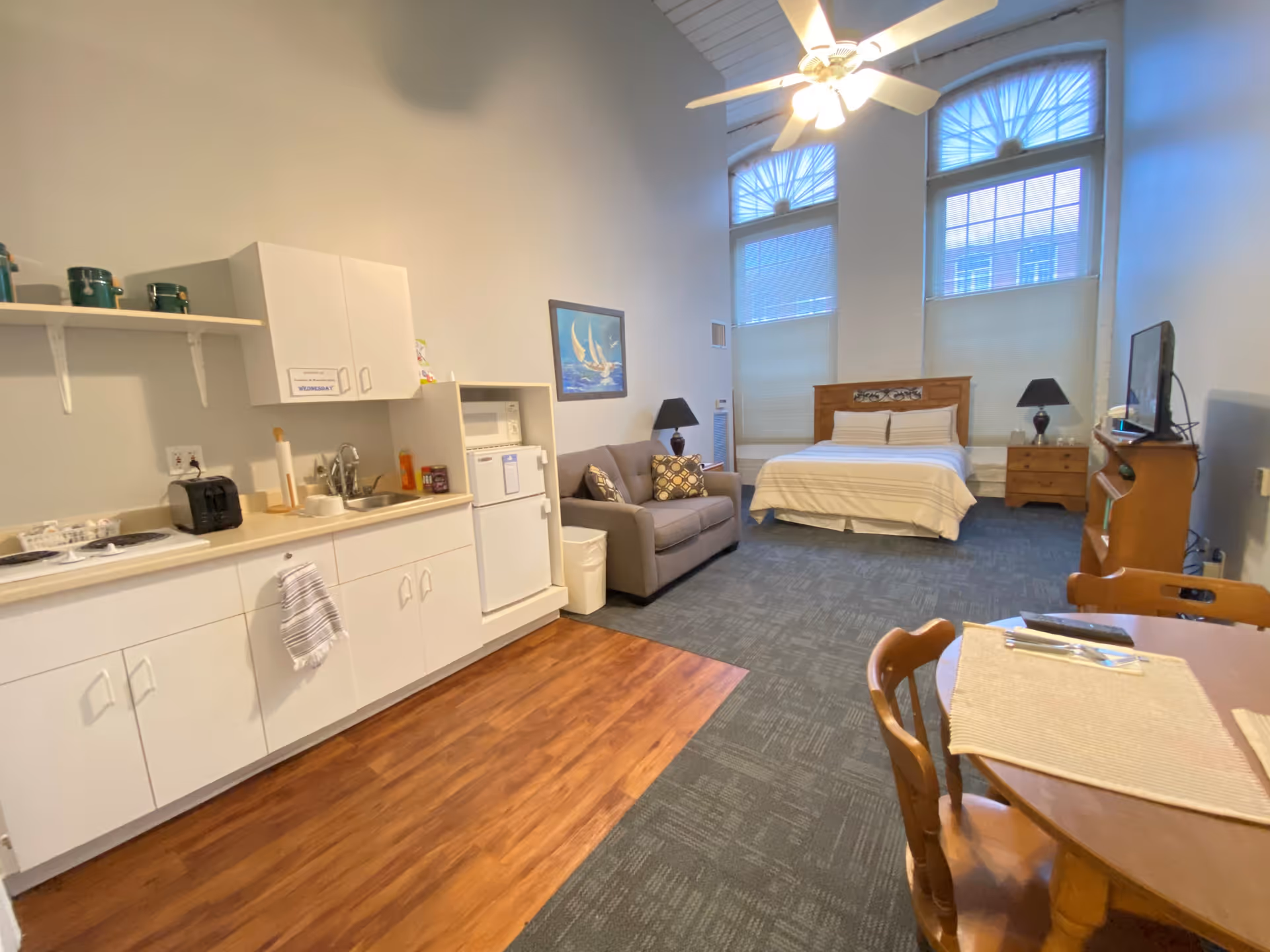 A spacious assisted living studio apartment featuring a kitchenette with white cabinets, a small refrigerator, microwave, and toaster. The room includes a gray loveseat, a wooden bed with white bedding, two nightstands with lamps, a wooden dining table with chairs, and a TV on a wooden stand. Large windows with blinds and a ceiling fan provide natural light and ventilation.