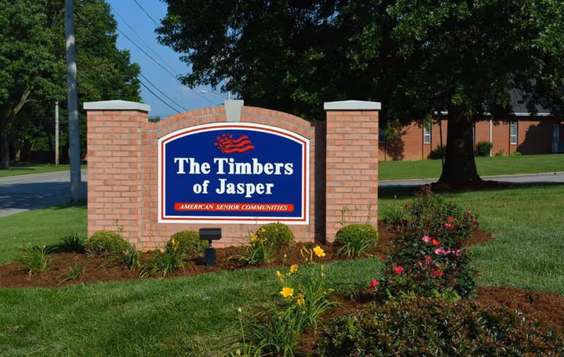 Brick entrance sign reading "The Timbers of Jasper" set in landscaped grass and flowers with trees and a building behind.