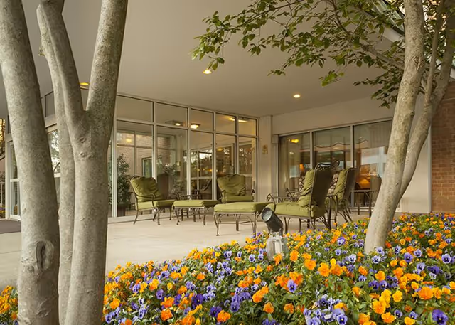 Outdoor patio area at Bedford Court with green cushioned chairs and tables on a concrete floor, surrounded by colorful orange, yellow, and purple flowers and trees.