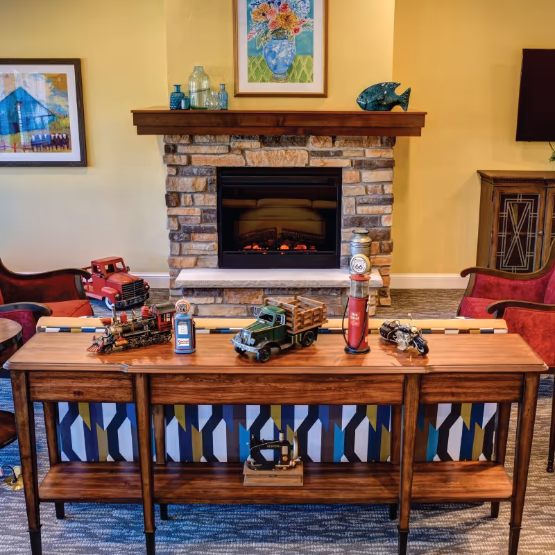 A cozy living room area featuring a stone fireplace with a wooden mantel. On the mantel are decorative glass bottles and a ceramic fish. Above the mantel hangs a colorful floral painting. In front of the fireplace is a wooden console table displaying vintage toy vehicles and gas pumps. The room has red upholstered chairs on either side and a patterned carpet.