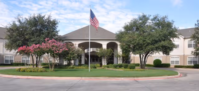 Front exterior view of a senior living facility with a circular driveway, an American flag on a flagpole in the center, and trees and shrubs surrounding the entrance under a partly cloudy sky.