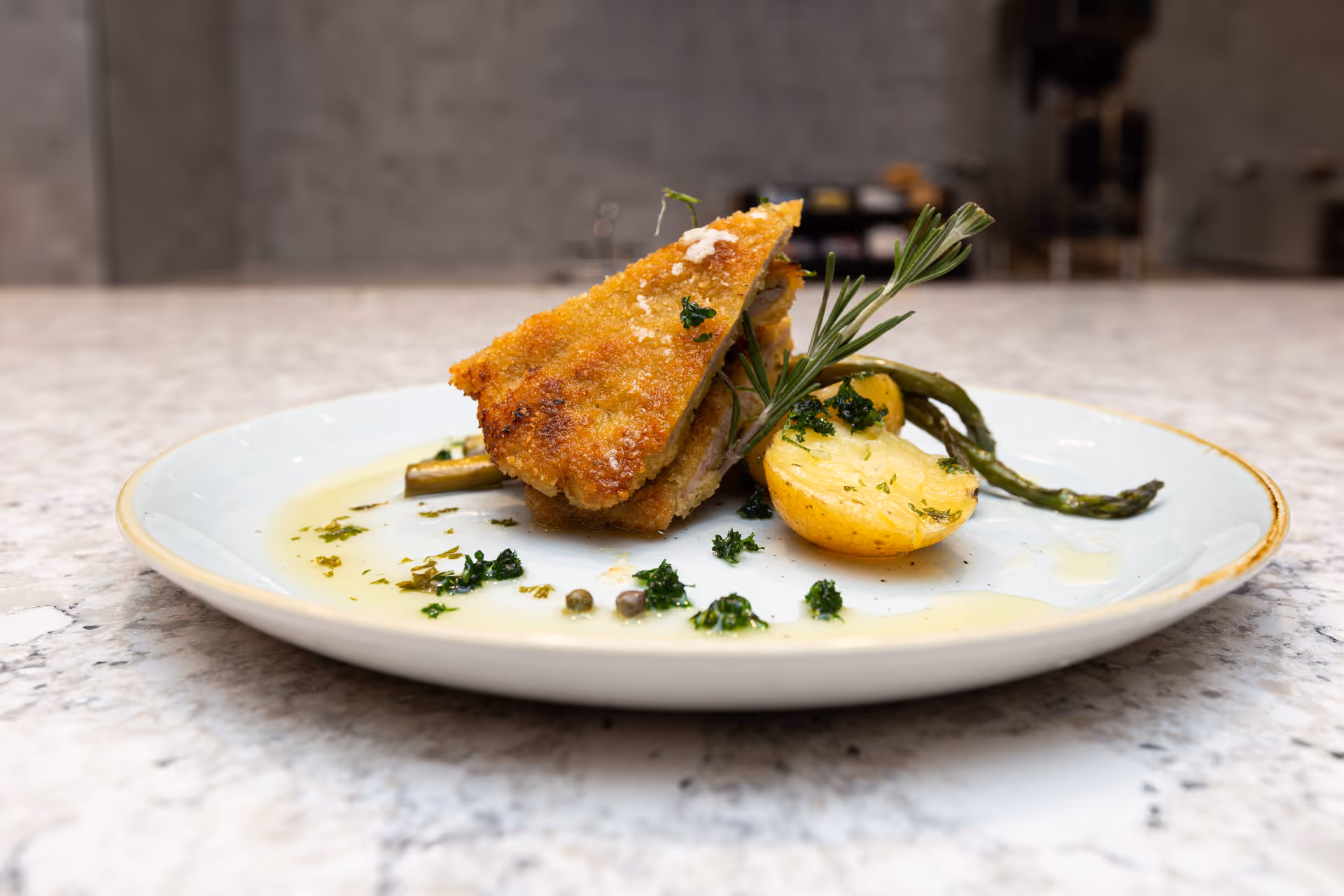 A plate with a breaded and fried piece of meat, garnished with a sprig of rosemary, served with roasted potatoes and asparagus, drizzled with a light sauce and sprinkled with herbs.