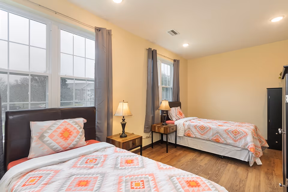 A bright bedroom with two twin beds featuring matching geometric patterned bedding in shades of orange, pink, and gray. Each bed has a dark brown headboard and a wooden nightstand with a lamp. The room has light yellow walls, wood flooring, and two large windows with gray curtains letting in natural light.