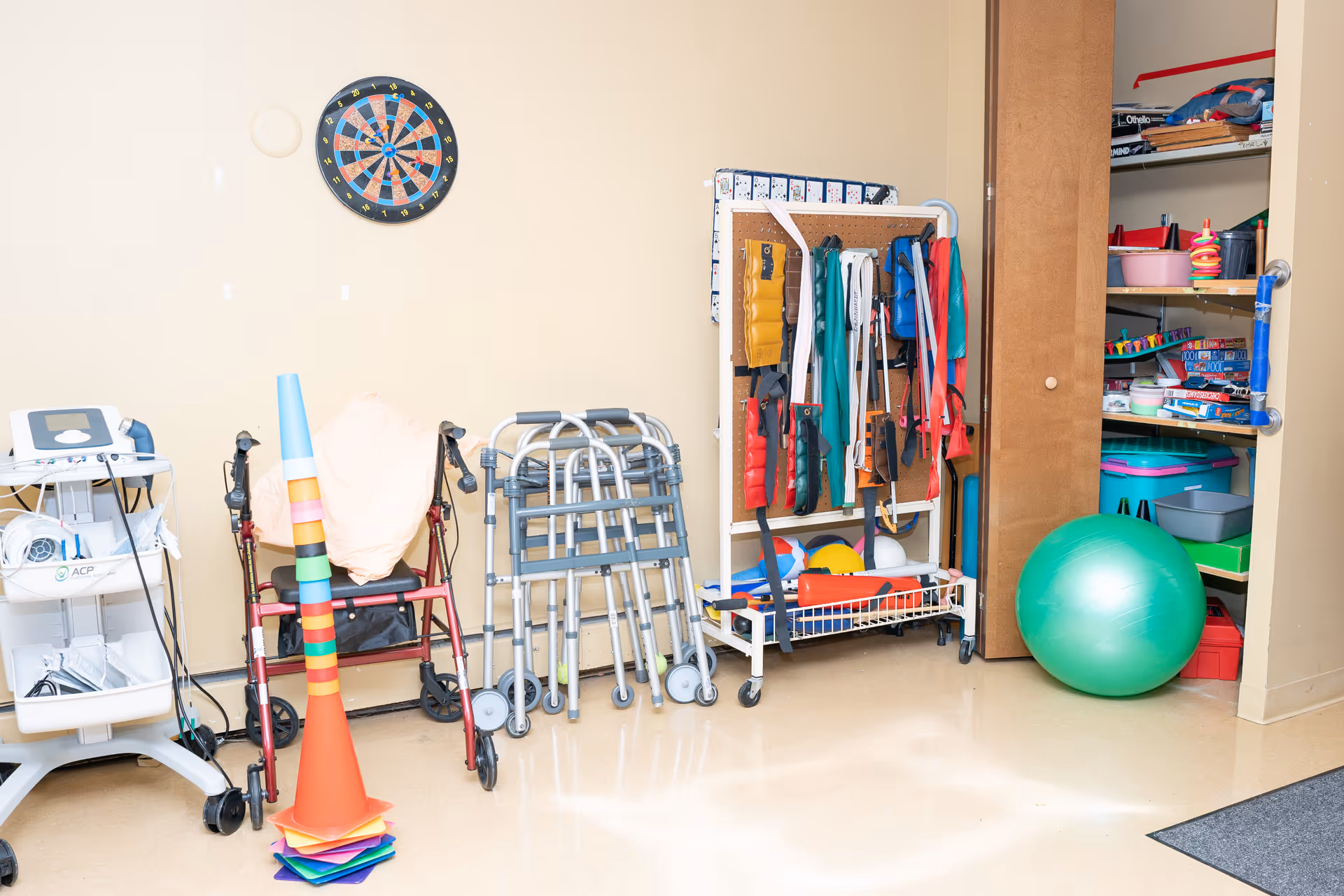 A therapy or rehabilitation room corner with various mobility aids including walkers and a red rollator, a stack of colorful cones, a dartboard on the wall, a rack holding exercise bands and other therapy equipment, and an open closet filled with games and a large green exercise ball.