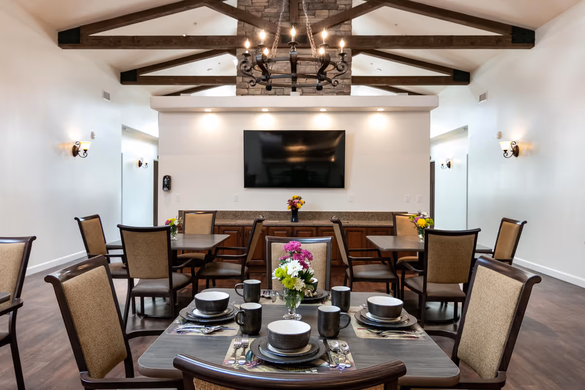 A dining room with several tables and chairs arranged neatly. Each table is set with plates, bowls, mugs, and silverware, and decorated with small flower arrangements. The room features a high ceiling with exposed wooden beams, a large flat-screen TV mounted on the far wall, and warm lighting from wall sconces and a chandelier.