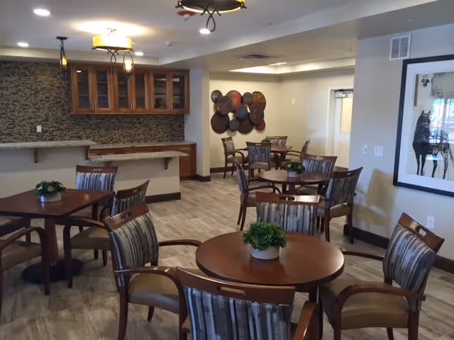 A well-lit dining area with several round wooden tables, each surrounded by four striped cushioned chairs. Small green potted plants are placed on each table. The room features a countertop with wooden cabinets above it, a decorative wall piece with circular elements, and a framed artwork on the wall. The flooring is light-colored wood, and the walls are painted in neutral tones.