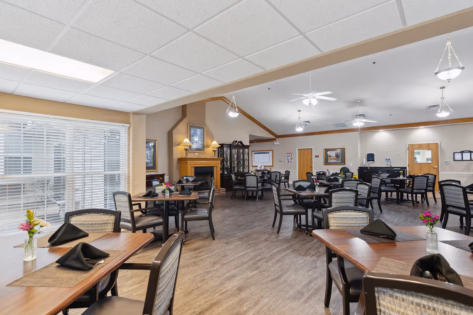 A spacious dining room with multiple wooden tables and chairs arranged neatly. Each table has a small vase with flowers and black folded napkins. The room features a fireplace with a wooden mantle, framed artwork on the walls, and large windows with white blinds allowing natural light to enter. The ceiling has recessed lighting and ceiling fans, and the floor is covered with wood-style flooring.