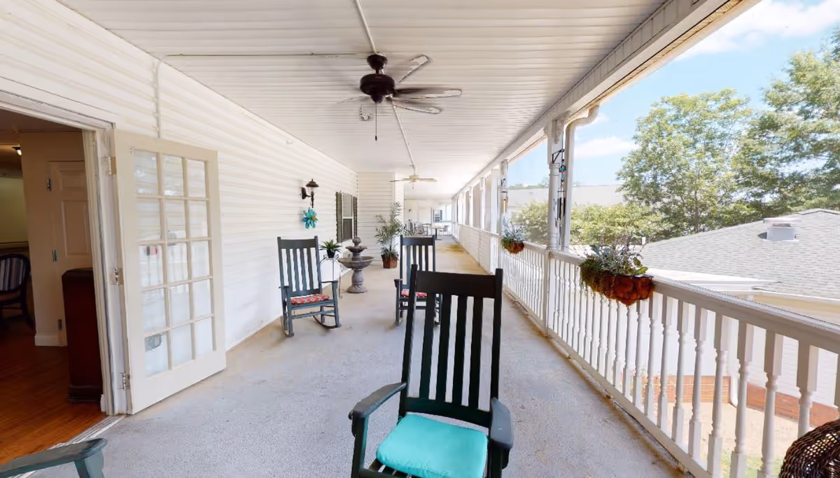 Long covered porch with rocking chairs, ceiling fans, potted plants, and a white railing overlooking trees and rooftops.