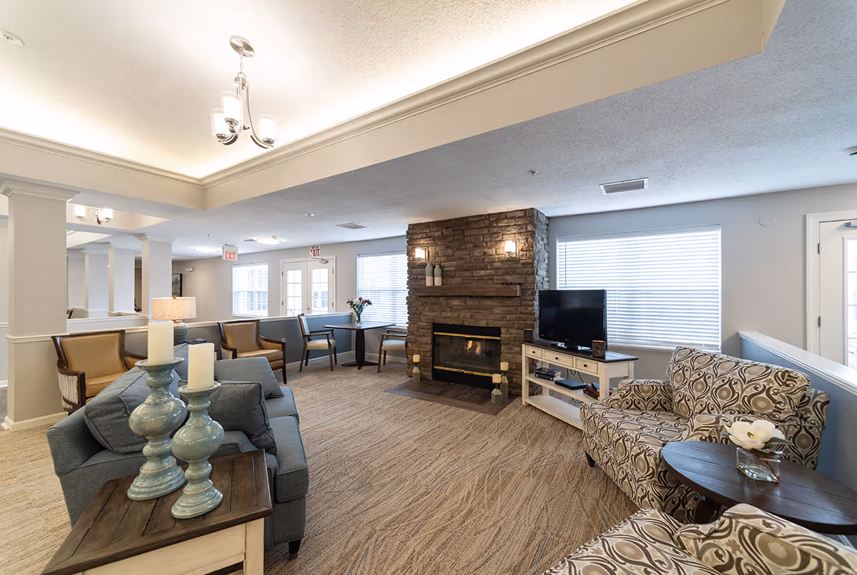 A cozy living room area in a senior living facility with a stone fireplace, a flat-screen TV on a white stand, patterned armchairs, a blue sofa, wooden side tables with decorative candles, and multiple chairs and tables near windows with blinds. The room has beige carpeting, soft lighting, and neutral-colored walls.