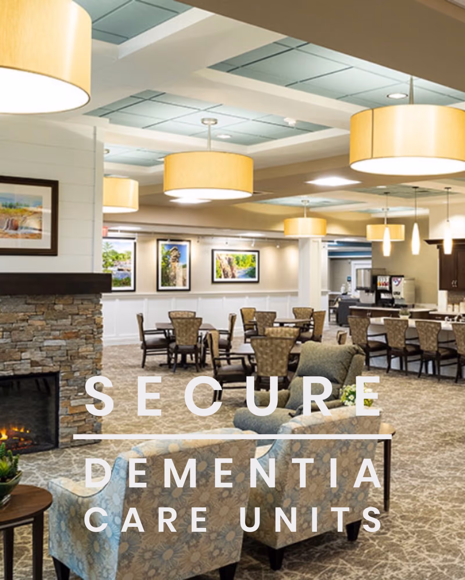 A spacious, well-lit common area with comfortable armchairs and dining tables. The room features a stone fireplace on the left, large hanging ceiling lights, framed landscape pictures on the walls, and a beverage station in the background.